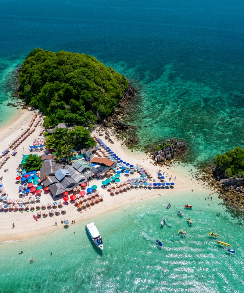 Vista aerea di una piccola isola tropicale con una spiaggia di sabbia bianca a forma di mezzaluna, bagnata da acque cristalline turchesi. Sulla spiaggia si vedono ombrelloni colorati, lettini e un bar, mentre diverse persone nuotano o fanno kayak nelle acque circostanti