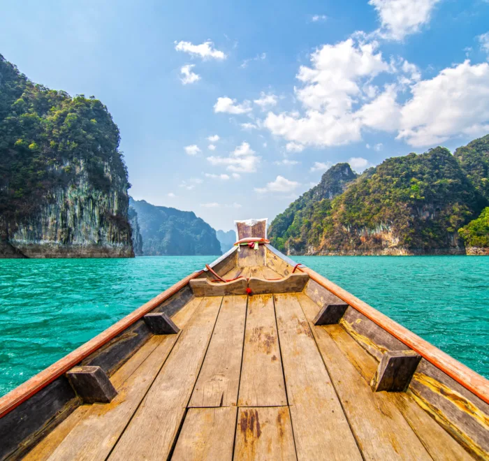 Vista dalla prua di una tradizionale barca in legno che naviga su acque turchesi e cristalline, circondata da imponenti scogliere calcaree ricoperte da una fitta giungla tropicale. Il cielo è di un azzurro intenso con nuvole bianche e gonfie, evocando la maestosa bellezza del Parco Nazionale di Khao Sok o di un paesaggio simile