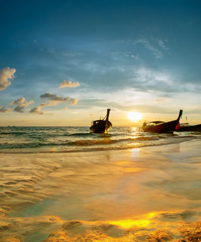 Tramonto mozzafiato su una spiaggia tropicale, con il sole che cala sull'orizzonte illuminando il cielo nuvoloso e due longtail boat silhouette che fluttuano nell'acqua bassa e dorata