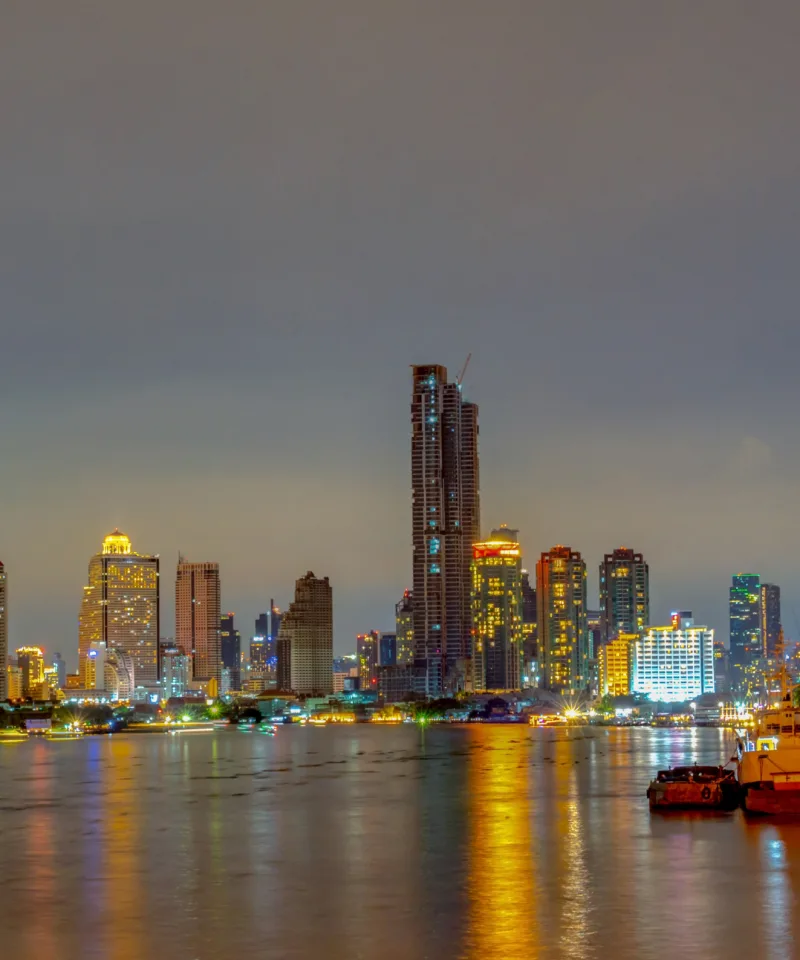 Vista notturna del lungofiume di una grande città, con un fiume che riflette le luci scintillanti dei numerosi grattacieli e degli edifici moderni che si ergono lungo la riva. Una grande ruota panoramica illuminata è visibile sulla destra, e alcune barche sono ormeggiate sulle acque scure