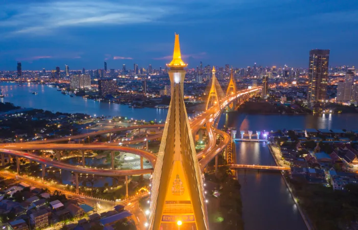 Vista aerea notturna del ponte Bhumibol di Bangkok illuminato, con le sue imponenti torri dorate che si stagliano contro lo skyline cittadino e il fiume Chao Phraya