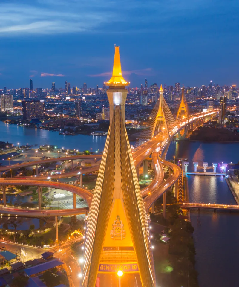 Vista aerea notturna del ponte Bhumibol di Bangkok illuminato, con le sue imponenti torri dorate che si stagliano contro lo skyline cittadino e il fiume Chao Phraya