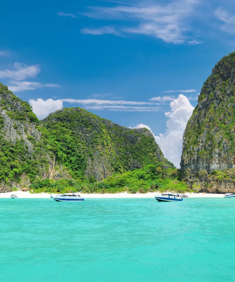 La famosa Maya Bay con scogliere carsiche verdeggianti che si ergono dalle acque cristalline e turchesi dell'oceano, con piccole imbarcazioni ormeggiate vicino alla spiaggia di sabbia bianca