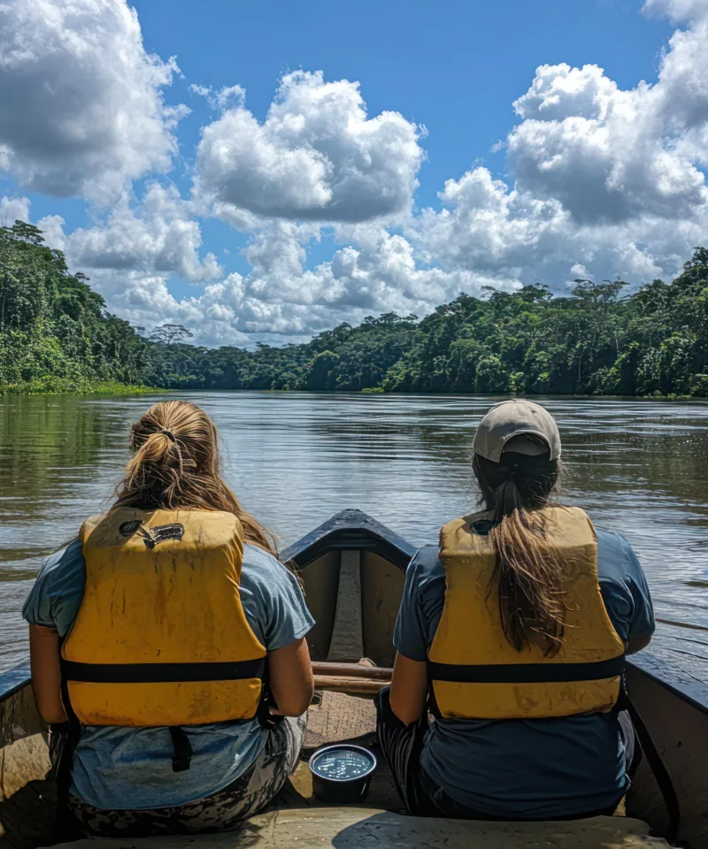 Due persone con giubbotti di salvataggio gialli navigano in una canoa su un fiume calmo, circondato dalla fitta e lussureggiante foresta pluviale. Il cielo è azzurro con nuvole bianche e gonfie che si riflettono sull'acqua, suggerendo un'avventura nella natura selvaggia