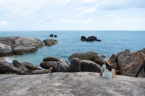 Una persona (vista di schiena) con un cappello di paglia e un vestito chiaro è seduta su un'ampia formazione rocciosa grigia, ammirando il mare blu e il cielo nuvoloso. Diverse grandi rocce affiorano dall'acqua e dalla costa rocciosa, creando un paesaggio costiero sereno e naturale