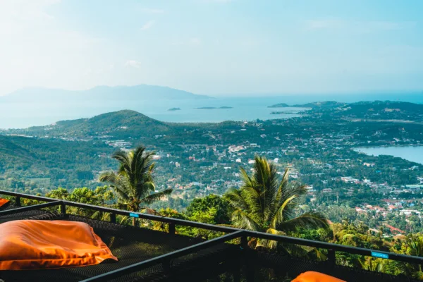 Veduta panoramica mozzafiato da un punto di osservazione elevato, con cuscini arancioni in primo piano su una terrazza. Il paesaggio si estende su una lussureggiante vegetazione tropicale e un'area urbana che si affaccia su un vasto tratto di mare e isole all'orizzonte sotto un cielo limpido. Palme in primo piano aggiungono un tocco esotico al panorama