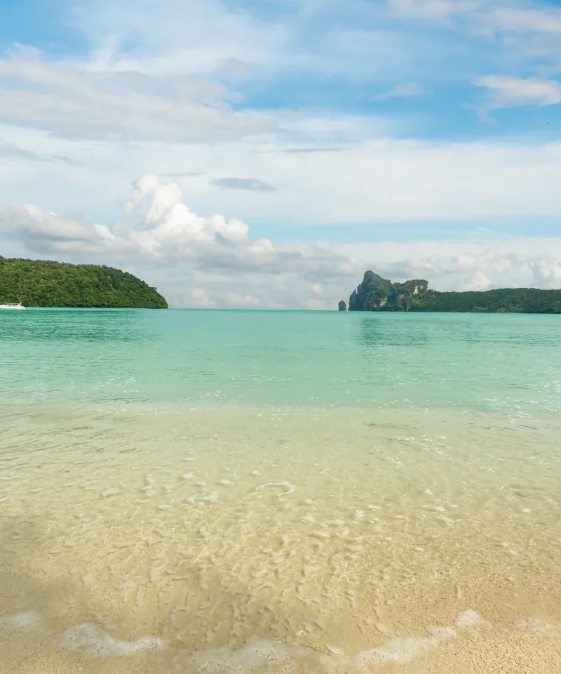 Acque turchesi e cristalline di una baia tropicale, con una spiaggia di sabbia chiara in primo piano e isole montuose verdi che si stagliano all'orizzonte sotto un cielo blu con nuvole sparse