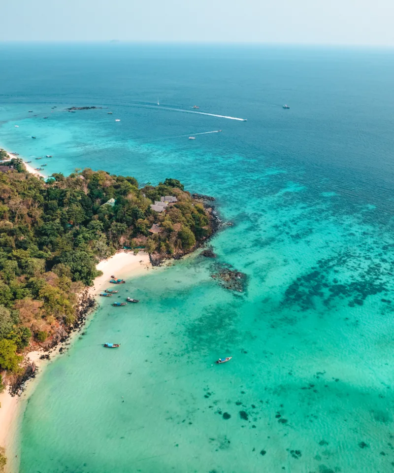 Vista aerea di una spiaggia di sabbia bianca bagnata da acque turchesi cristalline, con una fitta vegetazione tropicale che si estende fino alla costa. Diverse piccole imbarcazioni sono ancorate nell'acqua, mentre l'oceano aperto si estende all'orizzonte