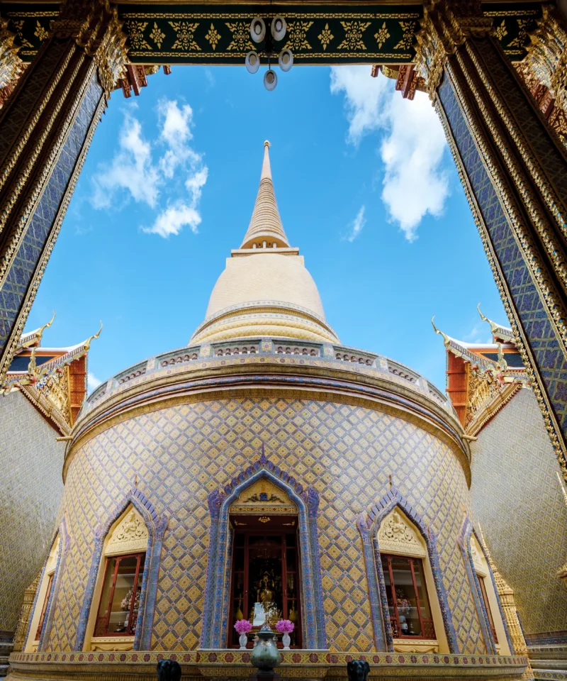 Un'imponente chedi dorata si erge su una base circolare decorata con piastrelle a motivi geometrici blu e oro, in un tempio buddista tailandese. La struttura è vista attraverso un portale riccamente decorato, sotto un cielo azzurro con nuvole sparse