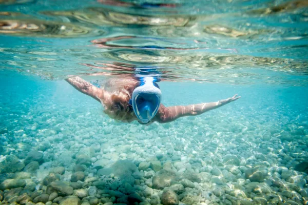 Una persona fa snorkeling sott'acqua, indossando una maschera integrale e pinne, mentre nuota sopra un fondale marino roccioso e ghiaioso. La superficie dell'acqua riflette la luce solare, creando un'atmosfera luminosa e chiara