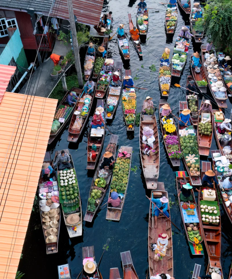 Vista aerea di un vivace mercato galleggiante, dove numerose barche tradizionali sono affollate in un canale stretto. Ogni barca è piena di frutta, verdura e altri prodotti freschi, con venditori e acquirenti che interagiscono. Ai lati del canale si vedono i tetti delle case locali e la vegetazione