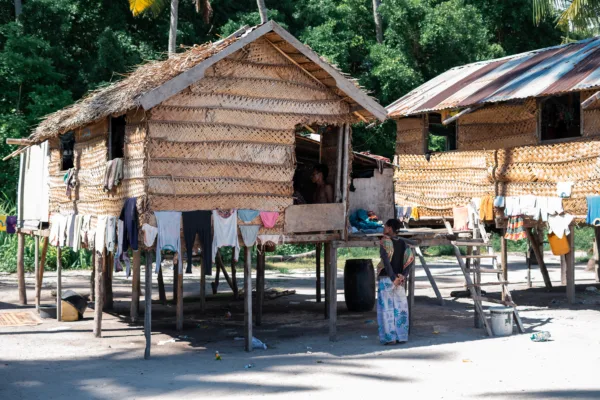 Case tradizionali su palafitte con tetti di paglia e pareti di bambù o foglie intrecciate in un villaggio tropicale, con vestiti stesi ad asciugare e una persona in piedi davanti a una delle abitazioni. Sullo sfondo si intravede una fitta vegetazione