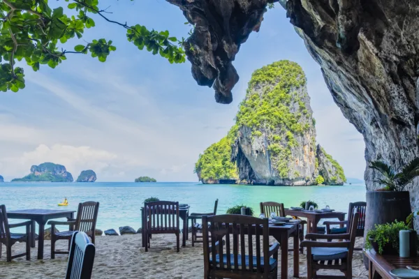 Tavoli e sedie di un ristorante all'aperto direttamente su una spiaggia sabbiosa, con una vista spettacolare su isole carsiche calcaree ricoperte di vegetazione che emergono dalle acque turchesi. La scena è incorniciata da una grotta o roccia a destra e foglie verdi in alto a sinistra