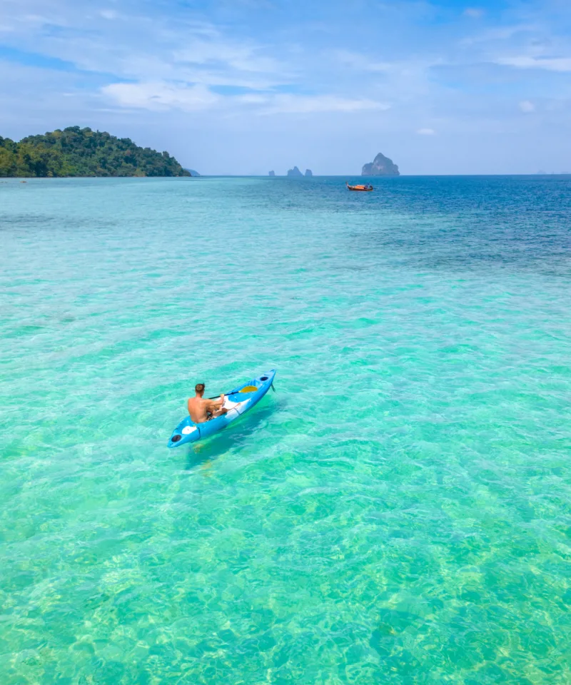 Un uomo rema su un kayak blu sulle acque cristalline e turchesi di un mare tropicale, con isole verdi e montagne carsiche in lontananza sotto un cielo blu