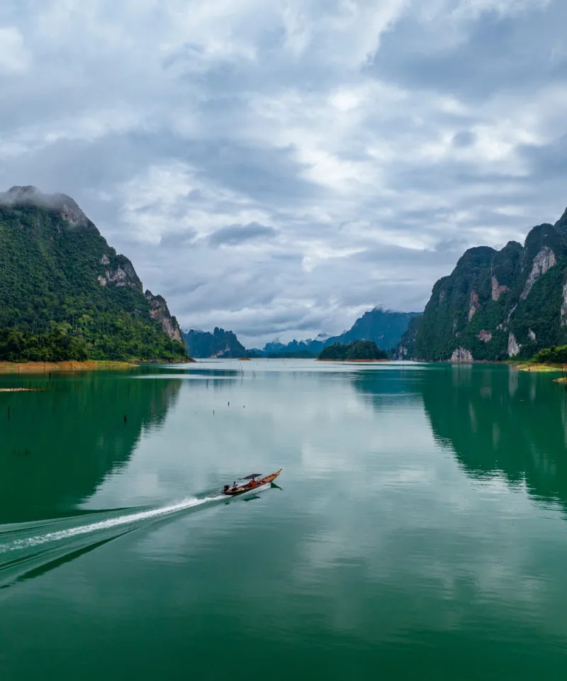 Ampia vista panoramica di un sereno lago di colore verde smeraldo, con una barca longtail che lascia una scia bianca sull'acqua, circondato da imponenti formazioni rocciose calcaree ricoperte di vegetazione lussureggiante e un cielo nuvoloso