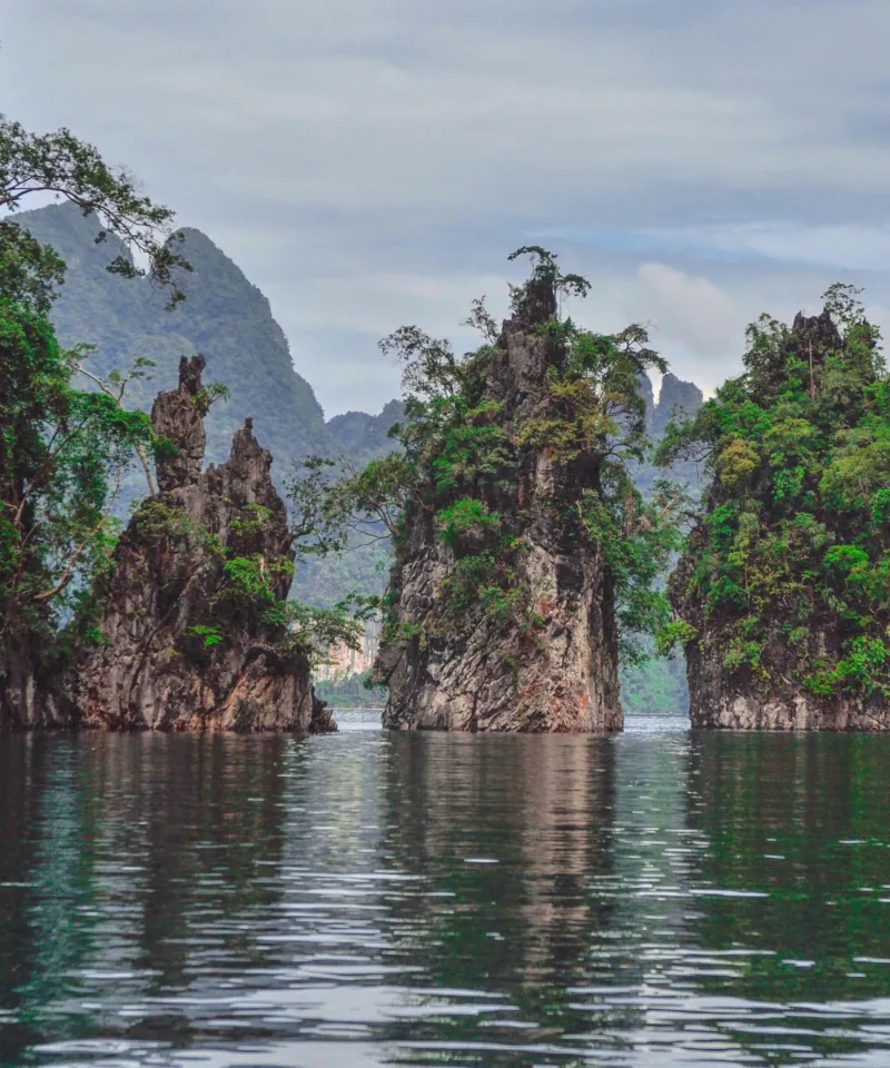 Tre imponenti formazioni rocciose calcaree coperte di vegetazione lussureggiante emergono dalle acque calme di un lago, con montagne sfocate sullo sfondo sotto un cielo nuvoloso