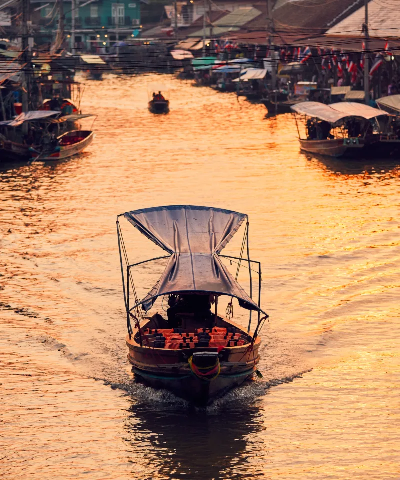 Una barca tradizionale con tettoia naviga su un fiume stretto e dorato al tramonto, lasciando una scia sull'acqua. Le sponde sono fiancheggiate da case in legno e altre barche, suggerendo un vivace mercato galleggiante
