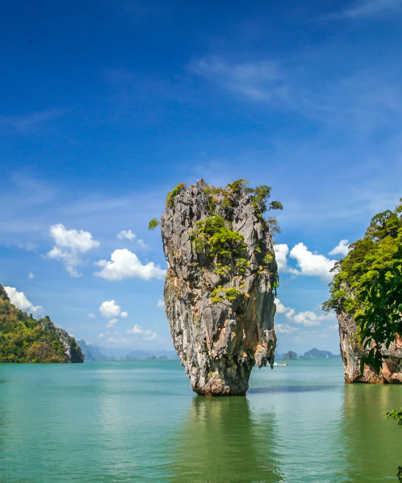 Una maestosa formazione rocciosa calcarea, conosciuta come Ko Tapu o 'James Bond Island', si erge verticalmente dalle acque verde smeraldo della Baia di Phang Nga. È circondata da altre isole coperte di vegetazione lussureggiante e un cielo azzurro con nuvole bianche sparse